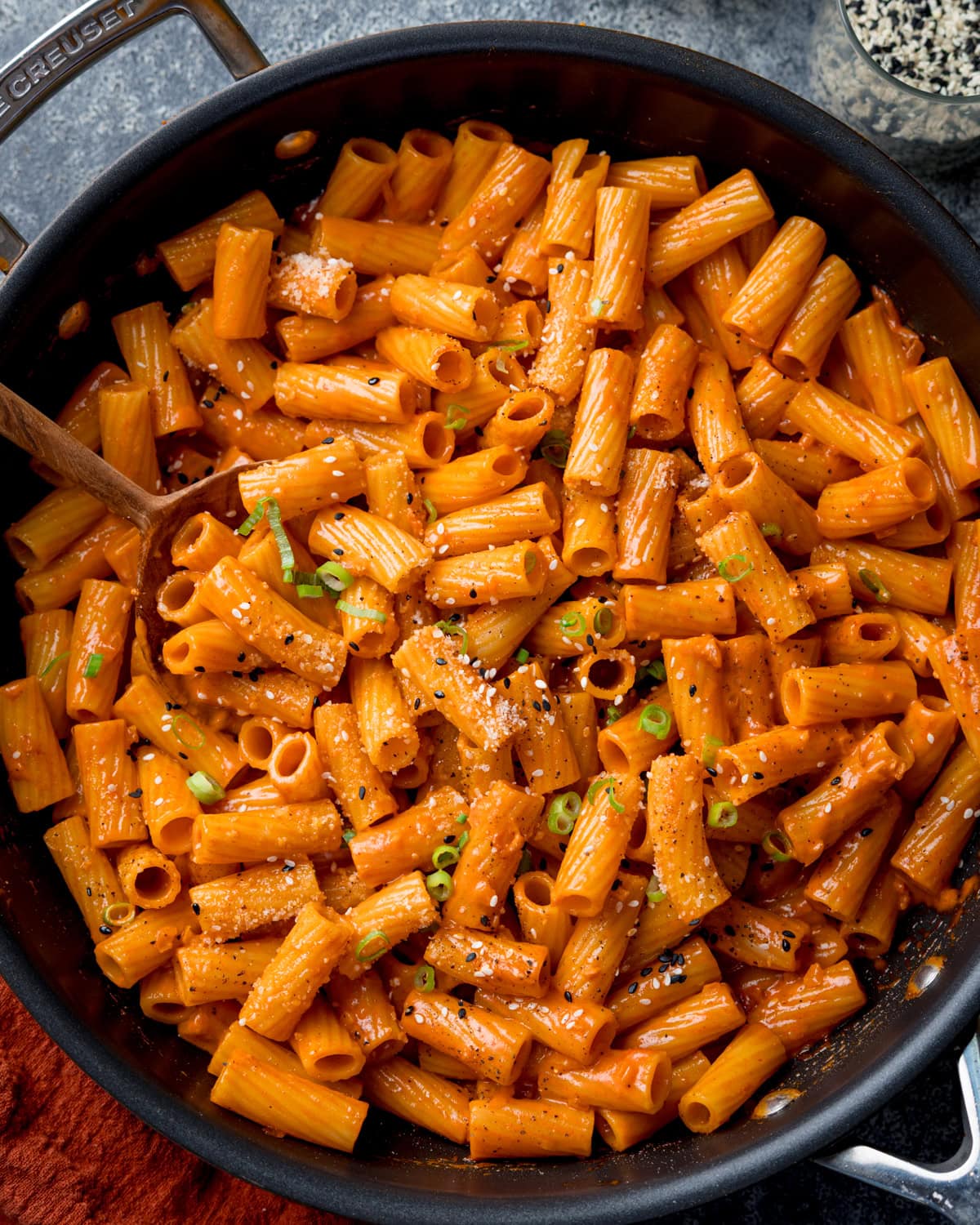 Overhead image of a frying pan filled with creamy Gochujang pasta topped with grated Parmesan, black pepper, and chopped spring onions. There is a wooden spoon resting in the pan.