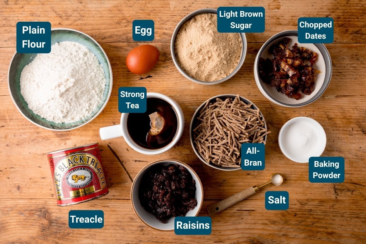 A wooden table ingredients for malt loaf. Ingredients are labelled, including plain flour, light brown sugar, All-Bran, chopped dates, raisins, baking powder, salt, an egg, a cup of strong tea and a tin of treacle.