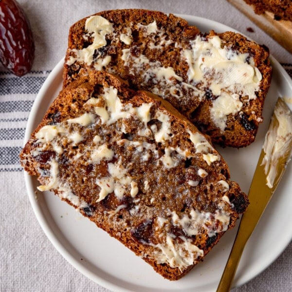 Two slices of malt loaf with butter spread on top, served on a white plate. A gold knife with butter on rests on the plate.