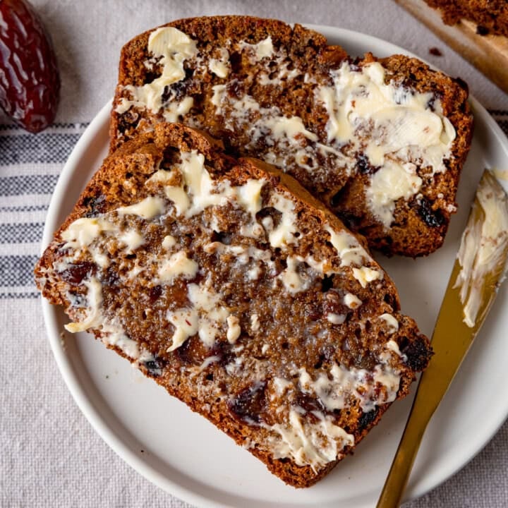 Two slices of malt loaf with butter spread on top, served on a white plate. A gold knife with butter on rests on the plate.