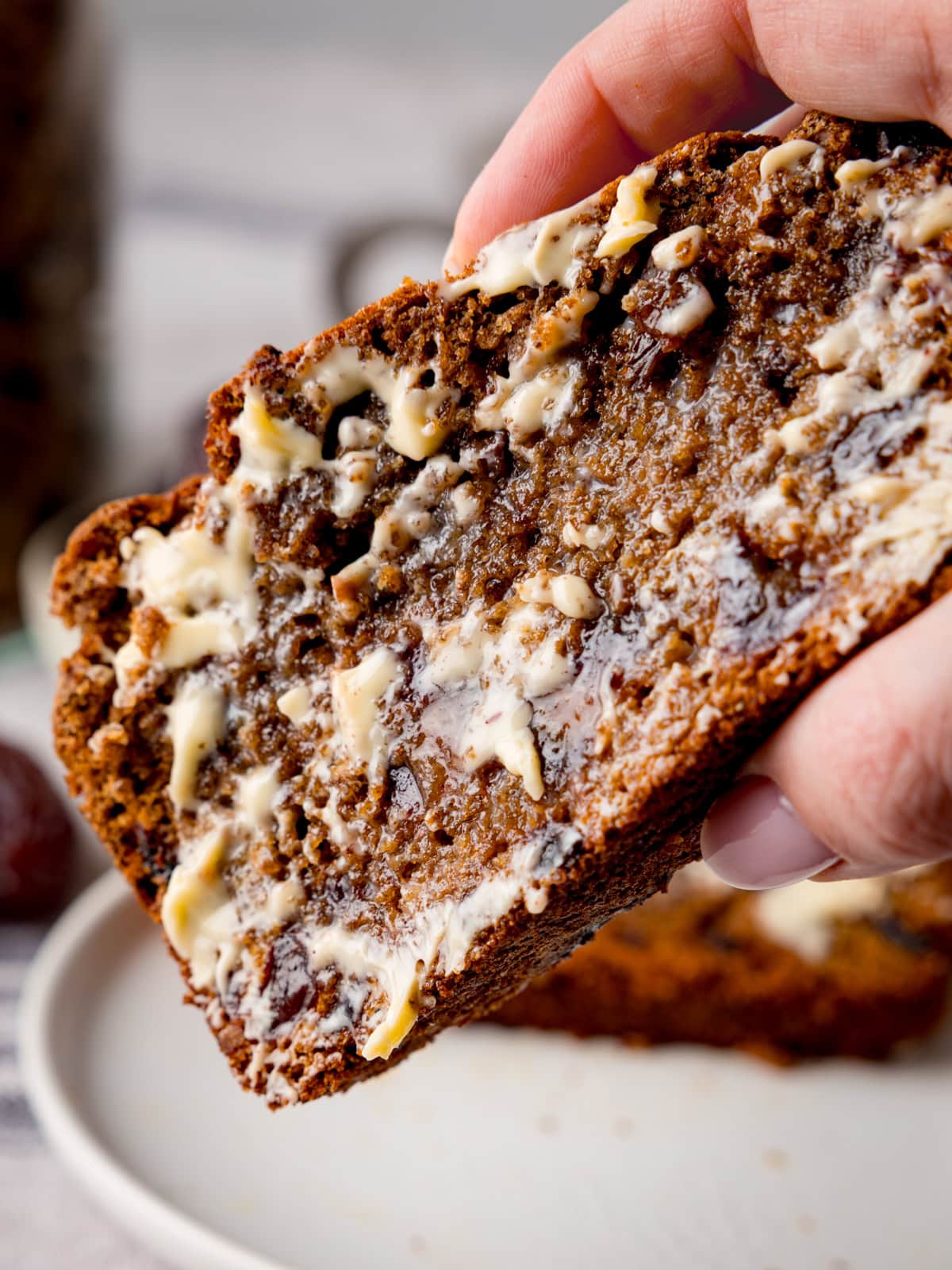 A hand holds a thick slice of buttered malt loaf, showing the bread&rsquo;s moist texture and just-melting butter.