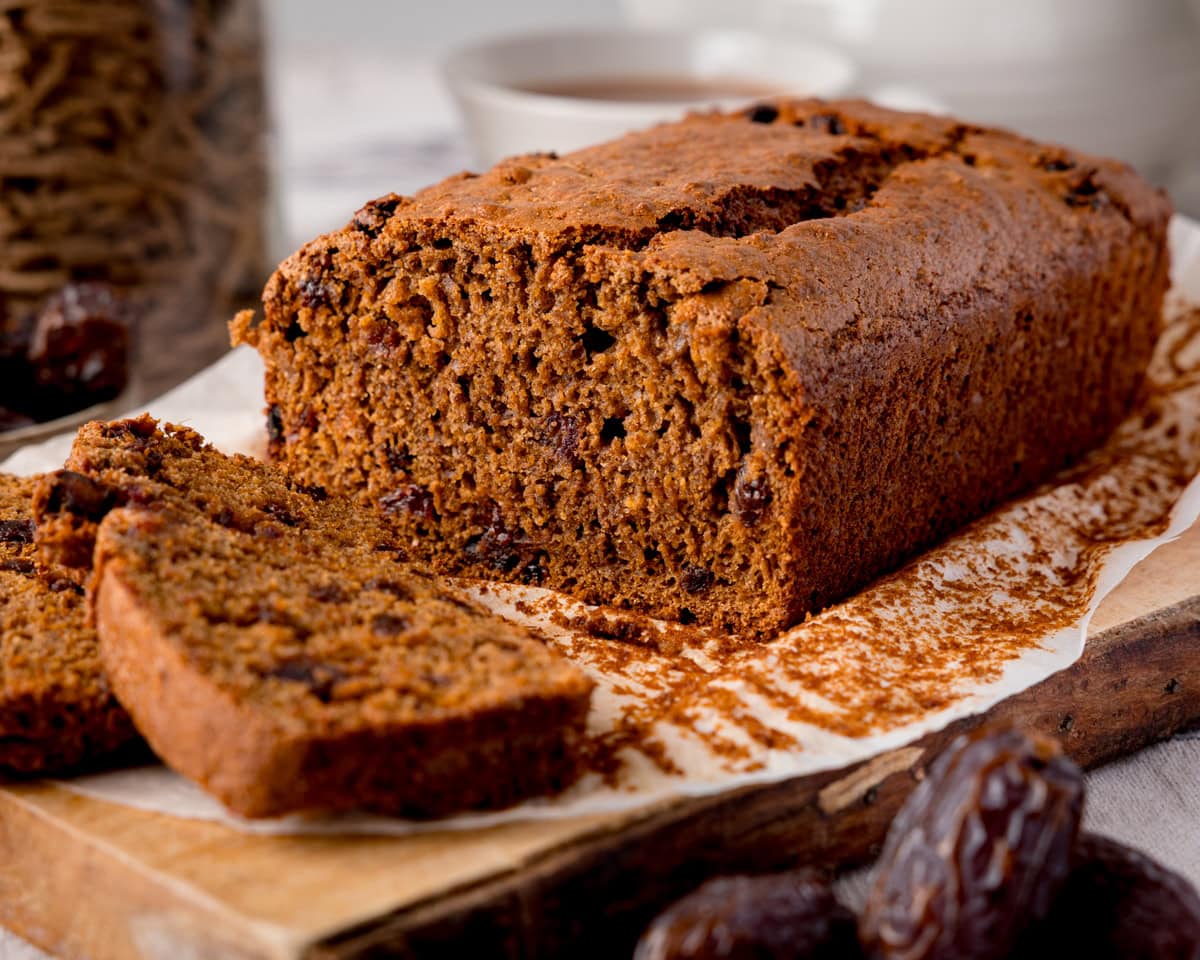 A loaf of sliced raising and date malt loaf sits on baking parchment, with a single slice cut and placed in front.
