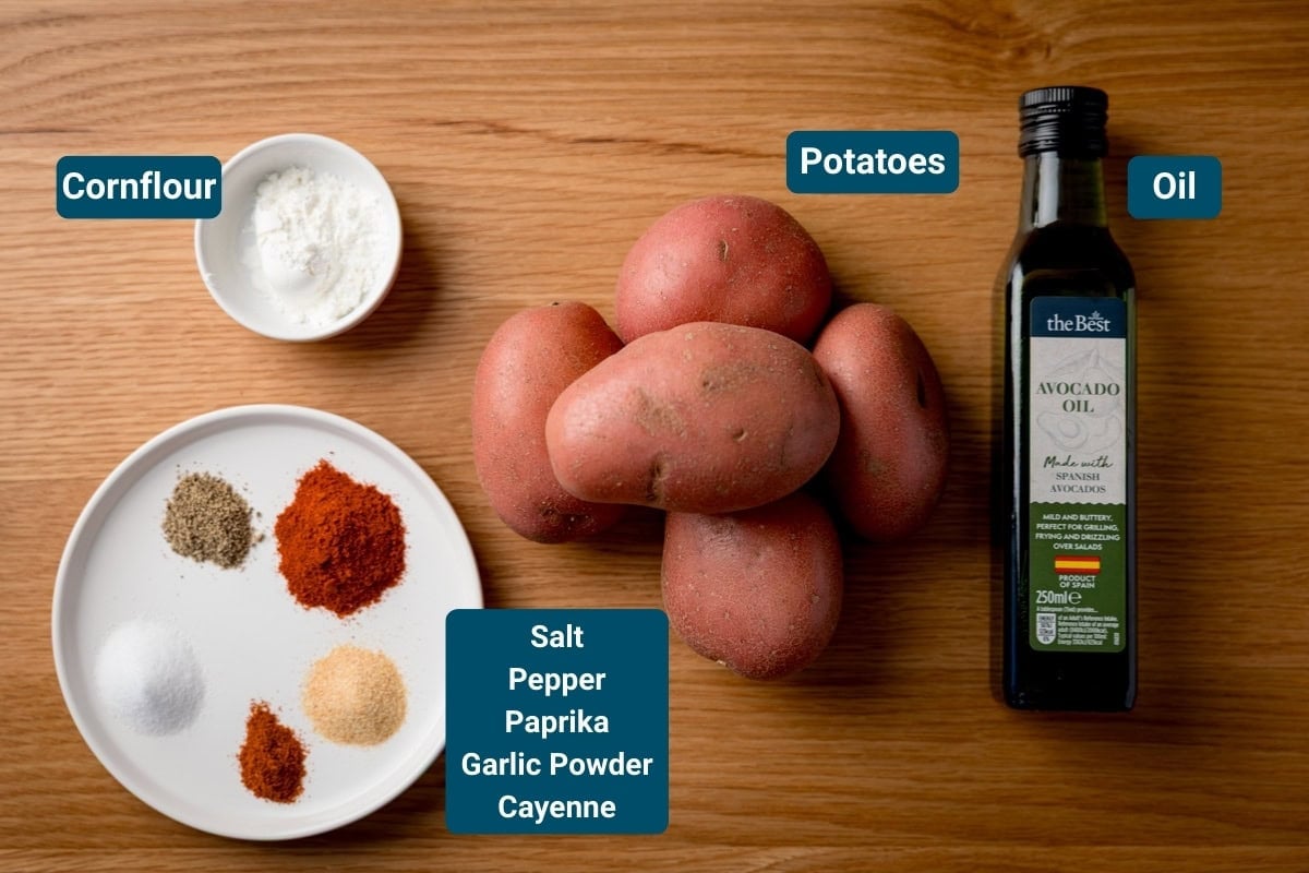 Ingredients for crispy potato wedges on a wooden surface. All ingredients are labelled, including: Cornflour, potatoes, a bottle of avocado oil and a plate with salt, pepper, paprika, garlic powder, and cayenne.