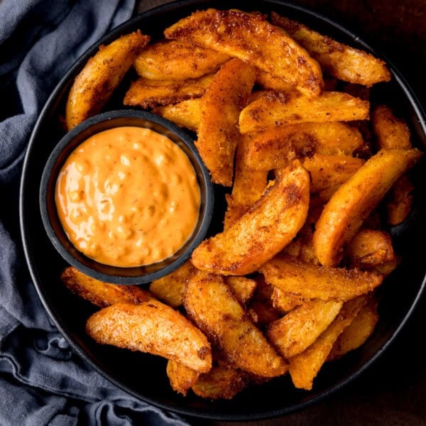 A black bowl filled with crispy potato wedges and a small dish of special sauce for dipping. The bowl is on a dark surface with a blue cloth nearby.