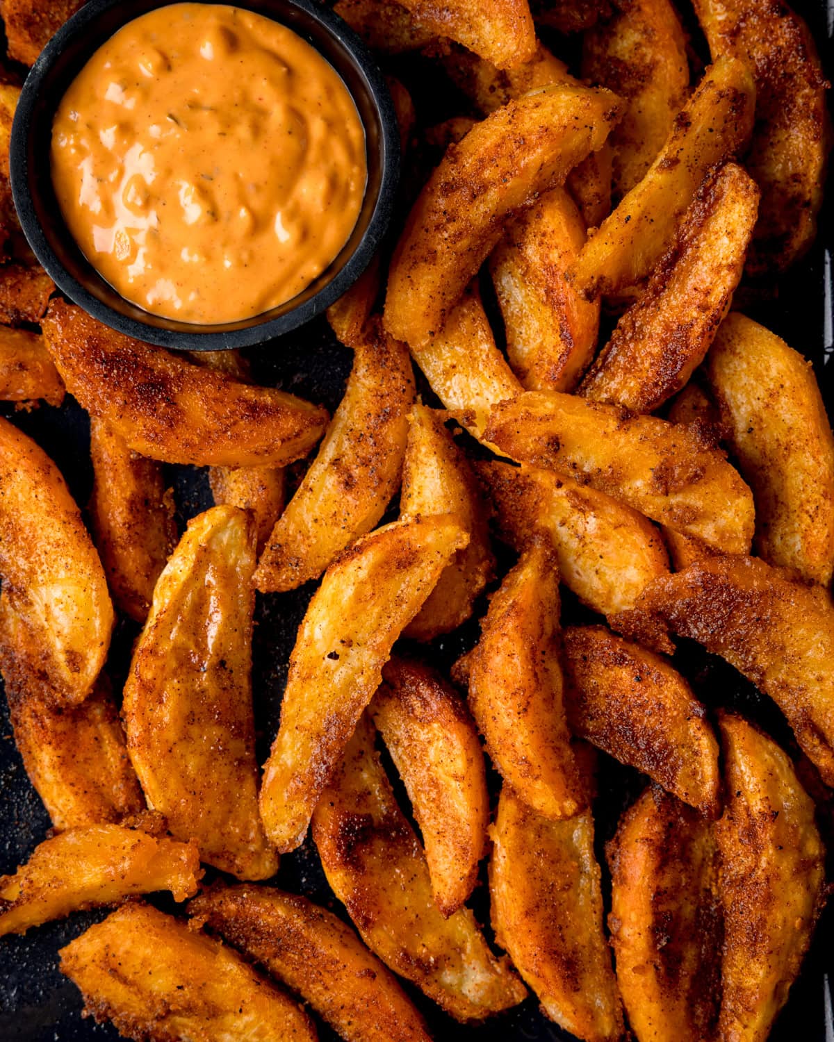 Close up overhead shot of crispy potato wedges on a dark baking tray. A small dish of special sauce for dipping is nestled into the wedges.