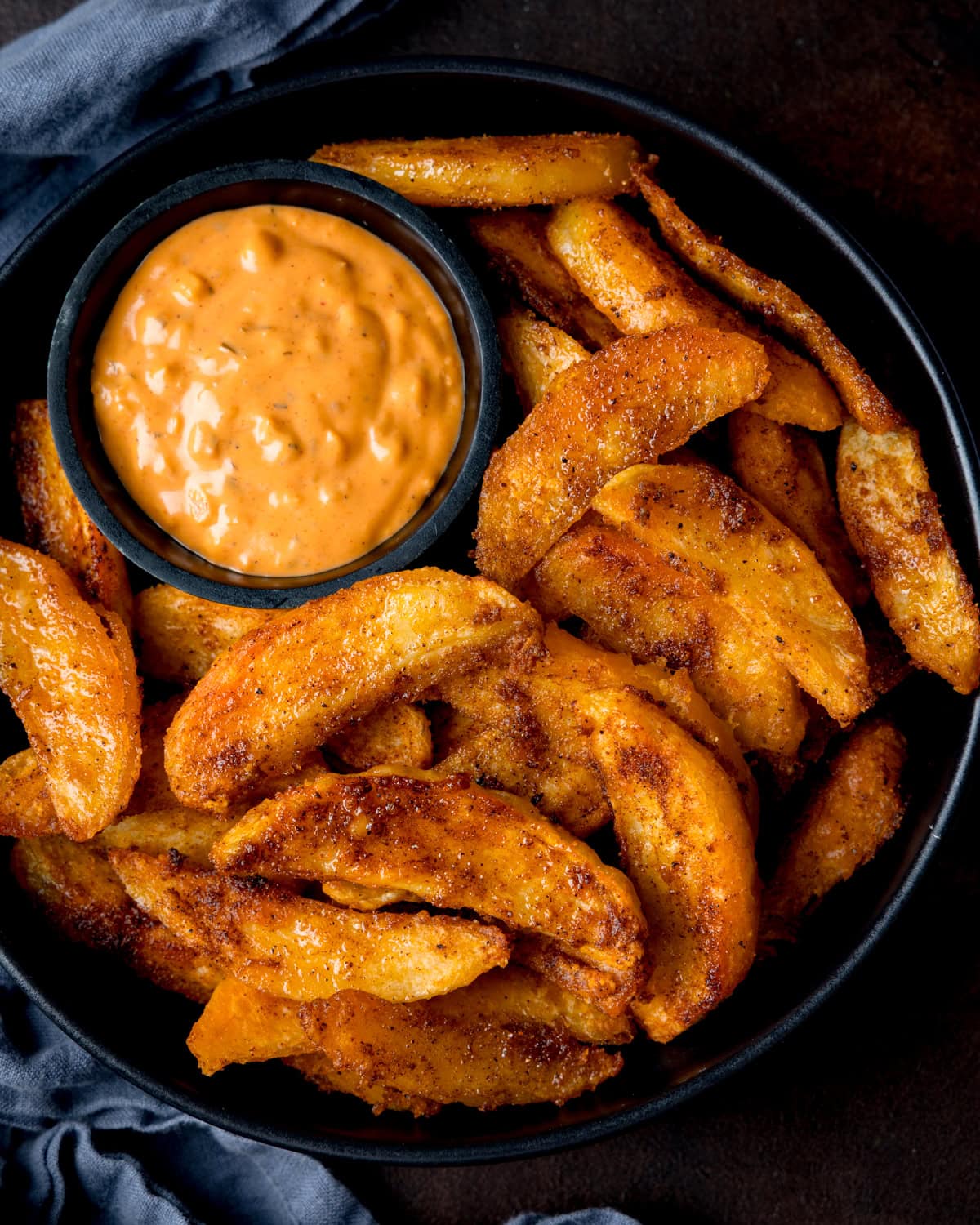 A black bowl filled with crispy potato wedges and a small dish of special sauce for dipping. The bowl is on a dark surface with a blue cloth nearby.