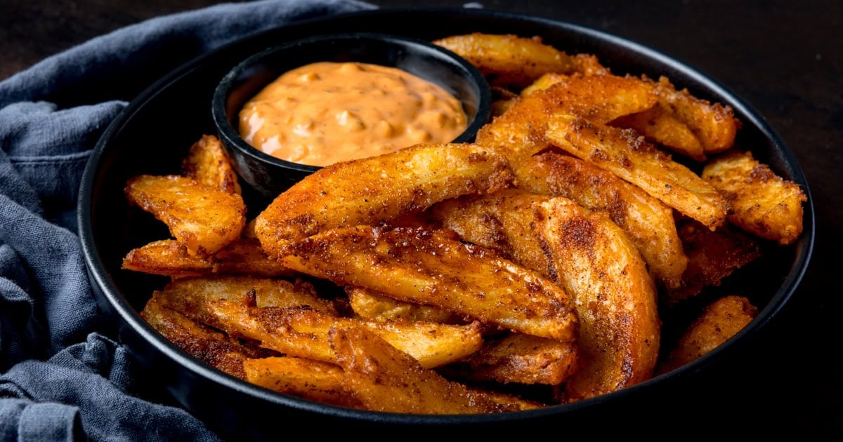 A black plate with crispy potato wedges and a small pot of creamy tomato dip.