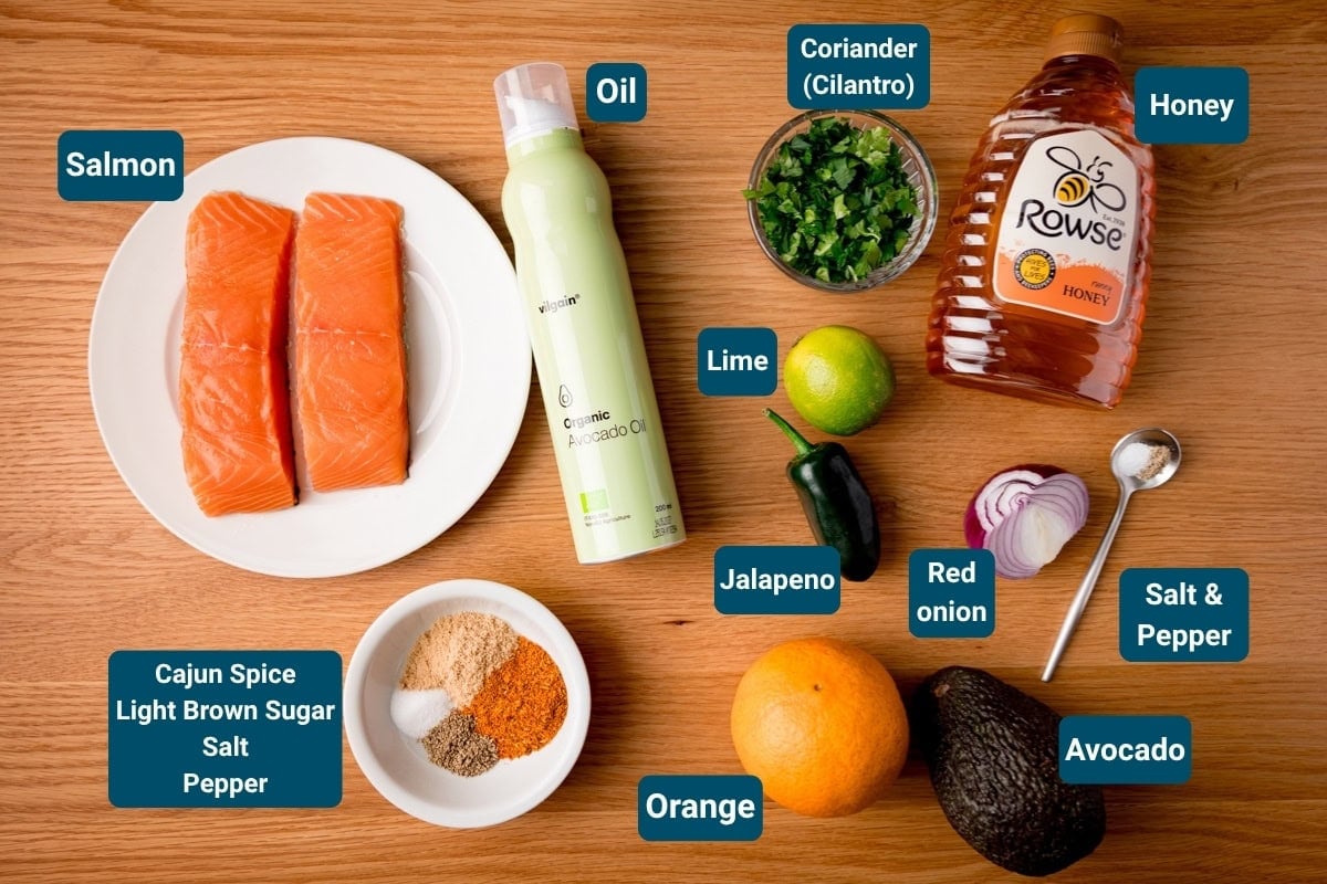 Ingredients for a salmon bowl recipe with orange-avocado salsa on a wooden surface. Includes: raw salmon fillets, avocado oil, honey, coriander, lime, jalape&ntilde;o, red onion, avocado orange salsa ingredients and spices. Each ingredient is labelled.