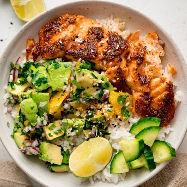 A white bowl on a white surface with spiced salmon, rice, avocado orange salad, cucumber, lime wedges, and a sprinkle of sesame seeds.