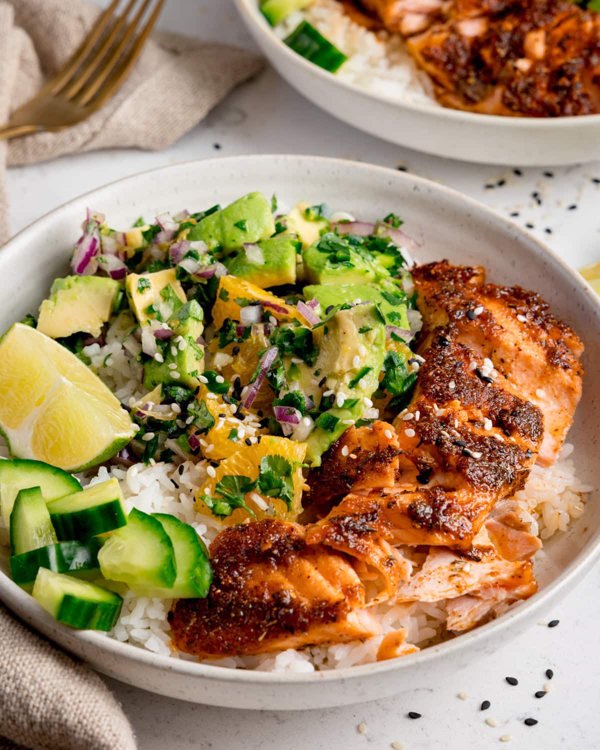 A salmon bowl with spiced air fryer salmon over white rice, topped with avocado orange salsa, cucumber pieces, coriander, and lime wedges.