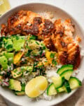 A white bowl on a white surface with spiced salmon, rice, avocado orange salad, cucumber, lime wedges, and a sprinkle of sesame seeds.