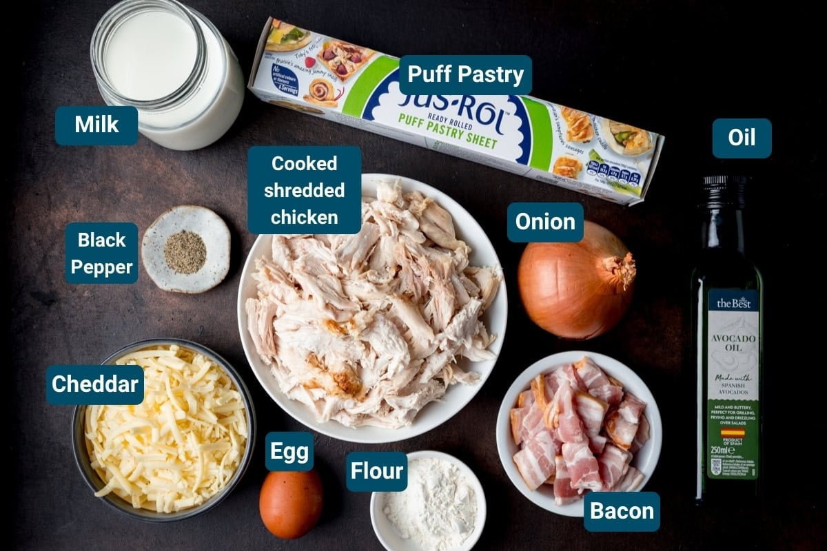 Ingredients for a cheesy chicken and bacon pasties - all labelled, including puff pastry, milk, black pepper, cheddar, egg, flour, cooked shredded chicken, onion, bacon and oil.