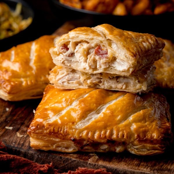 Square side-on image of a chicken and bacon pasty sliced in half with one piece stacked on the other to show the filling. There are two more pasties in shot, all on a wooden board. Sides of potato wedges and cabbage are in the background.