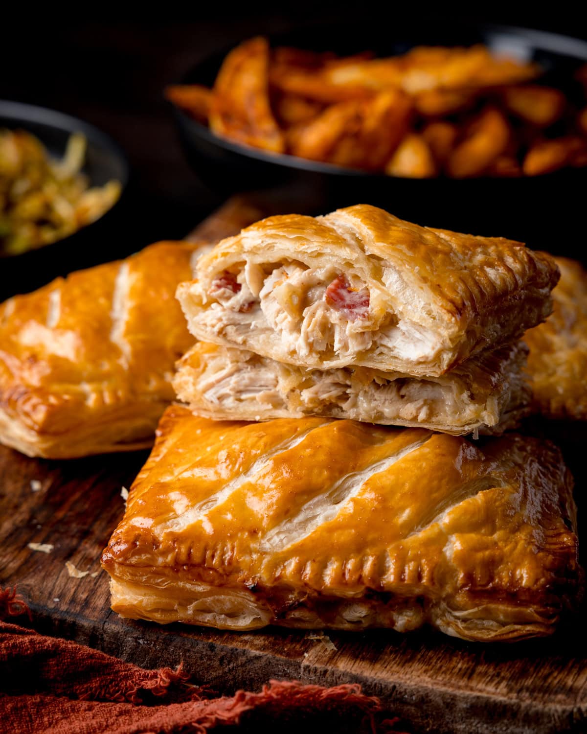 A chicken and bacon pasty sliced in half with one piece stacked on the other to show the filling. There are two more pasties in shot, all on a wooden board. Sides of potato wedges and cabbage are in the background.