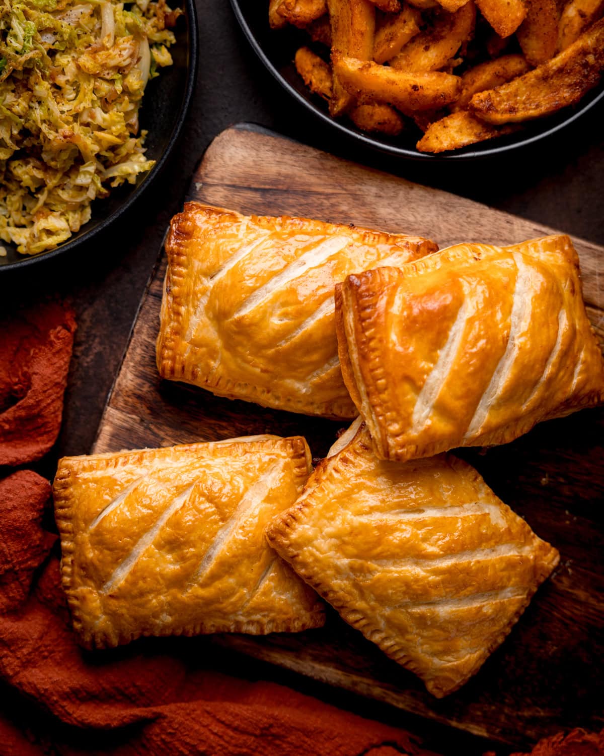 Four golden-brown cheesy chicken and bacon pasty bakes are arranged on a wooden board, with potato wedges and saut&eacute;ed cabbage in bowls nearby.