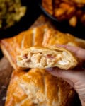 A hand holds half of a golden-brown Chicken and Bacon Pasty filled with shredded chicken and bacon, with other pastries and side dishes in the background.