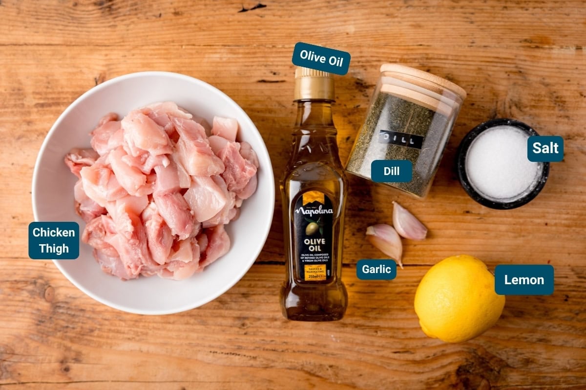 Labelled ingredients for chicken souvlaki on a wooden table, including: chicken thigh pieces, olive oil, dill, garlic cloves, salt, and a whole lemon.