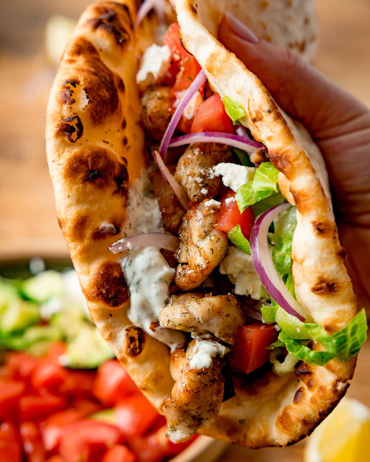 A hand holds a toasted flatbread filled with chicken souvlaki, lettuce, tomato, cucumber, red onion, and homemade tzatziki sauce. A bowl of salad is blurred in the background.