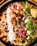 Overhead image of Chicken Souvlaki with tzatziki, lettuce, tomato, red onion and feta on toasted flatbread with a lemon wedge.