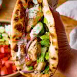 A hand holds a toasted flatbread filled with chicken souvlaki, lettuce, tomato, cucumber, red onion, and homemade tzatziki sauce.