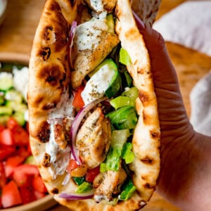 A hand holds a toasted flatbread filled with chicken souvlaki, lettuce, tomato, cucumber, red onion, and homemade tzatziki sauce.