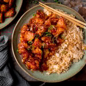 Overhead square image of a plate of fried rice and glazed Crispy Chilli Chicken topped with chopped spring onions, served with chopsticks.