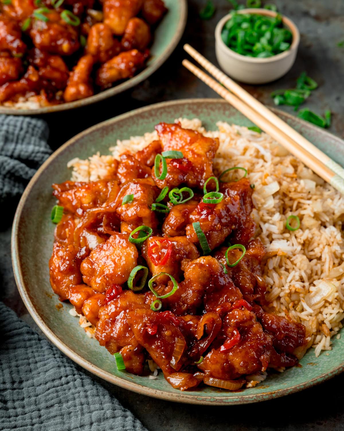 A plate of crispy sweet chilli chicken with sliced spring onions, served with rice and chopsticks. A bowl of spring onions and a further plate of chilli chicken is in the background.