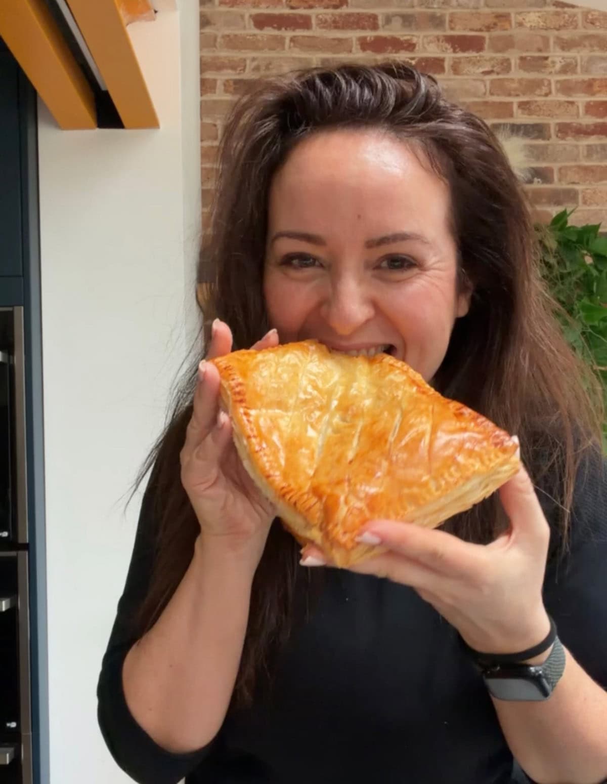 Nicky smiles as she takes a bite from a large cheesy chicken pasty, holding it with both hands..