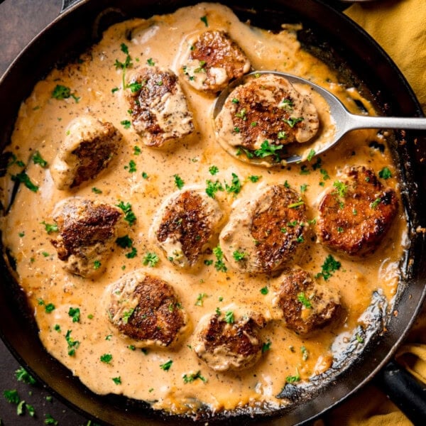 Pan of seared pork fillet medallions in a creamy Dijon sauce, garnished with chopped parsley, with a spoon lifting one piece.
