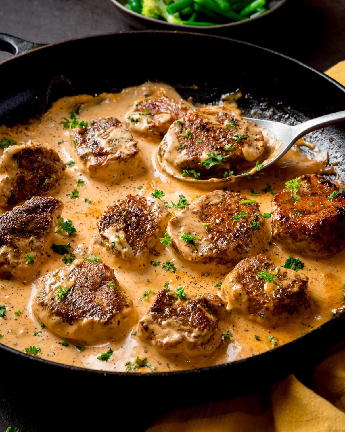 Pan-seared pork fillet medallions in a creamy Dijon sauce, garnished with chopped parsley, served in a black frying pan. There is a spoon lifting one piece.