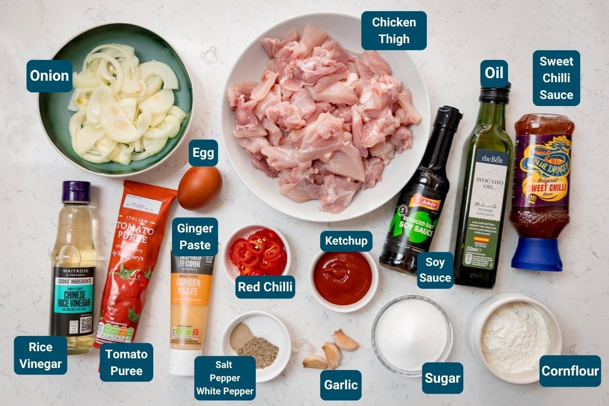 Ingredients for Crispy Chilli Chicken laid out on a white background. Each ingredient is labelled, including: chopped chicken thigh, sliced onion, sauces, spices, egg, garlic, red chilli, ginger paste and cornflour.