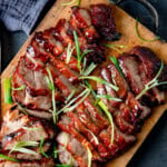 Sliced glazed char siu pork garnished with thin strips of spring onion, served on a wooden chopping board.
