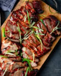 Sliced char siu pork with a glossy glaze, garnished with thinly sliced spring onions, served on a wooden chopping board.