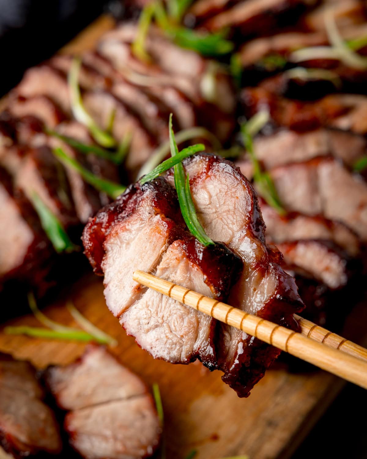 Chopsticks holding two slices of Chinese barbeque pork garnished with spring onions, more char siu on a wooden board in the background.