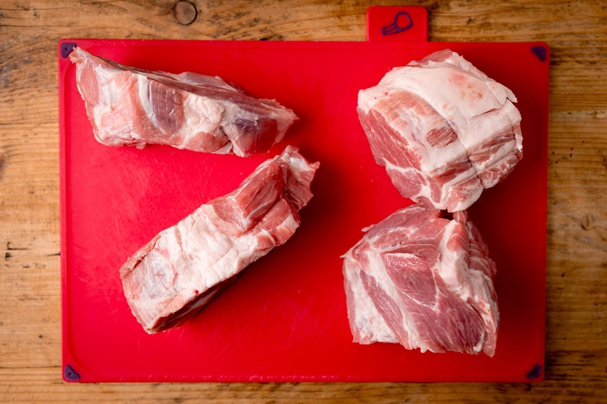Four pork shoulder cuts placed on a red chopping board atop a wooden surface.