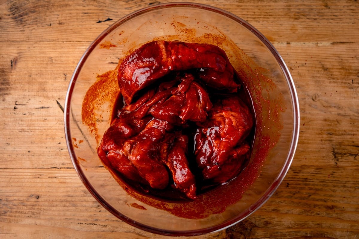 A glass bowl filled with raw char siu pork, coated in a red marinade. The bowl is on a wooden worktop.