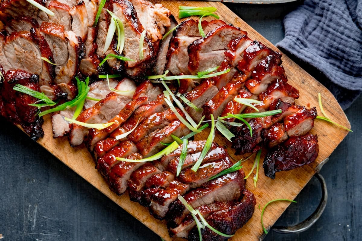 Wide image of sliced Chinese BBQ pork with a glossy red glaze is arranged on a wooden board and garnished with finely sliced spring onions.