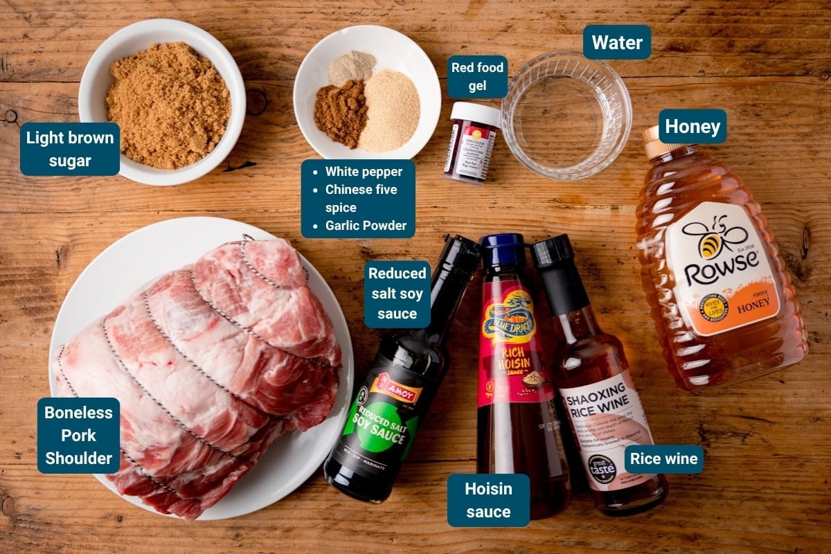 Ingredients for a char siu pork displayed on a wooden surface, including boneless pork shoulder, sauces, spices, sugar, water, honey, and red food colouring gel. Labels identify each item.