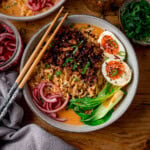 A bowl of spicy pork coconut curry ramen with sliced boiled eggs, pak choi, pickled onions and spring onions. There are a set of chopsticks resting o the bowl.
