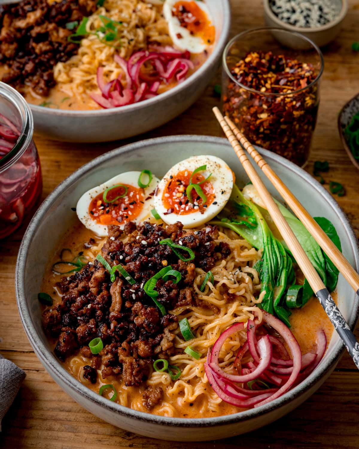 Two bowls of Curry Ramen with Spicy Pork, noodles, soft-boiled eggs, pak choi, pickled onions, and spring onions are served with chopsticks on a wooden table.