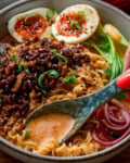 Close up of a bowl of Curry Ramen with Spicy Pork, halved soft-boiled eggs, pak choi, pickled onions, spring onions, and rich broth. A spoon is in the bowl (held by a hand, mostly out of shot) to capture the sauce.