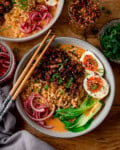 A bowl of spicy pork coconut curry ramen with sliced boiled eggs, pak choi, pickled onions and spring onions. There are a set of chopsticks resting o the bowl. A further bowl of ramen and small bowls of toppings are arranged around the main bowl, which sits on a wooden table.