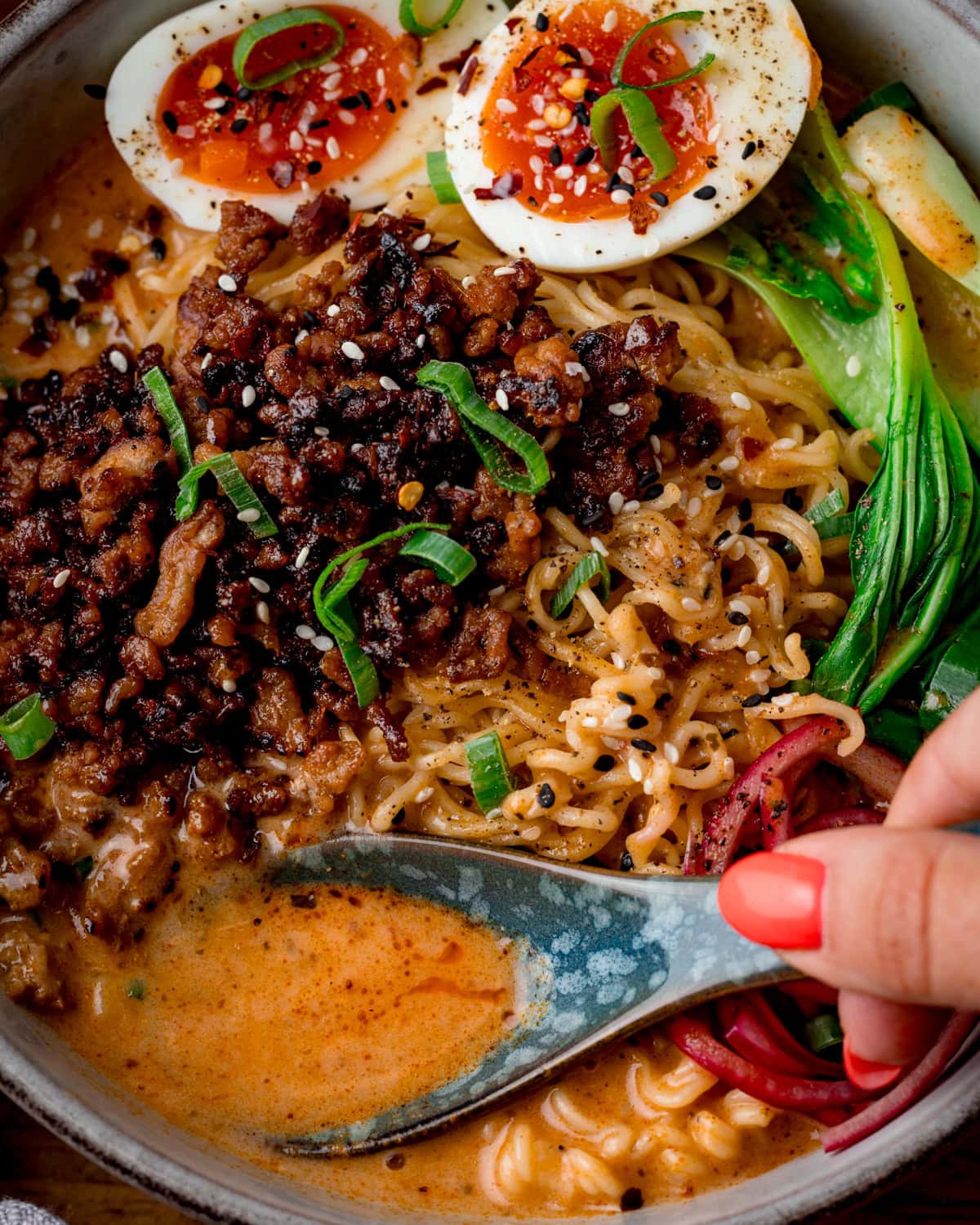 Close up of a bowl of Curry Ramen with Spicy Pork, halved soft-boiled eggs, pak choi, pickled onions, spring onions, and rich broth; a hand with red nails is holds a spoon in the bowl to capture the sauce.