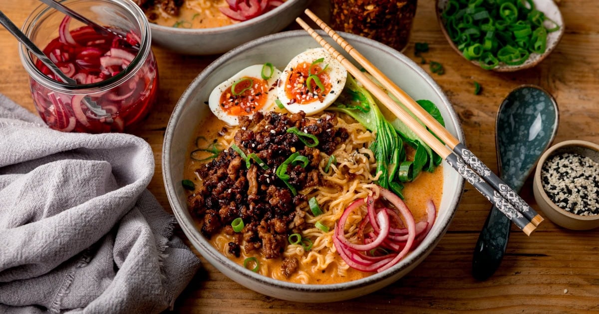 A round bowl with noodles in a broth, topped with crispy minced pork, jammy egg, red onion slices