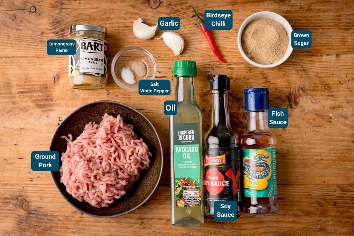 A top-down view of ingredients on a wooden table for spicy ground pork, including: pork mince, avocado oil, soy sauce, fish sauce, brown sugar, bird's eye chilli, garlic, lemongrass paste, salt, and white pepper. All ingredients are labelled.