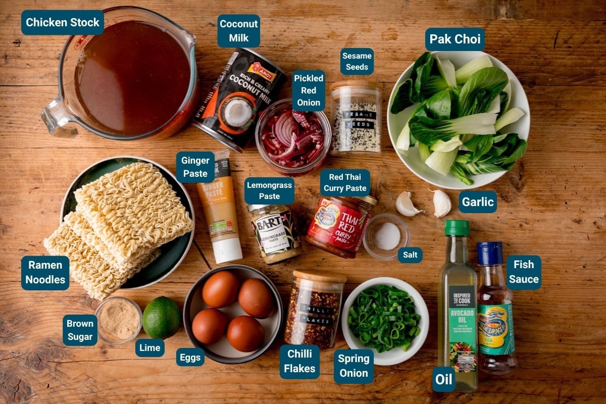 A top-down view of ingredients on a wooden table for curry ramen including chicken stock, coconut milk, pak choi, ramen noodles, eggs, lime, garlic and assorted sauces and seasonings. All ingredients are labelled.