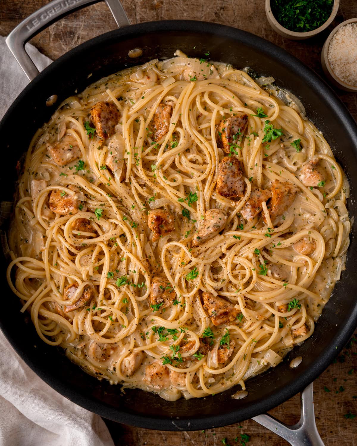 Overhead shot of a skillet filled with creamy Garlic Butter Chicken Pasta, pan-fried chicken pieces, chopped parsley, and a sprinkle of black pepper on top.