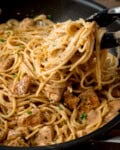 Tongs lifting creamy garlic butter chicken spaghetti with chunks of seasoned chicken and fresh parsley from a frying pan.