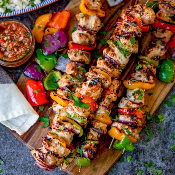 Grilled chicken and vegetable kebabs on a wooden board, garnished with herbs, with a bowl of salsa and assorted roasted vegetables on the side.