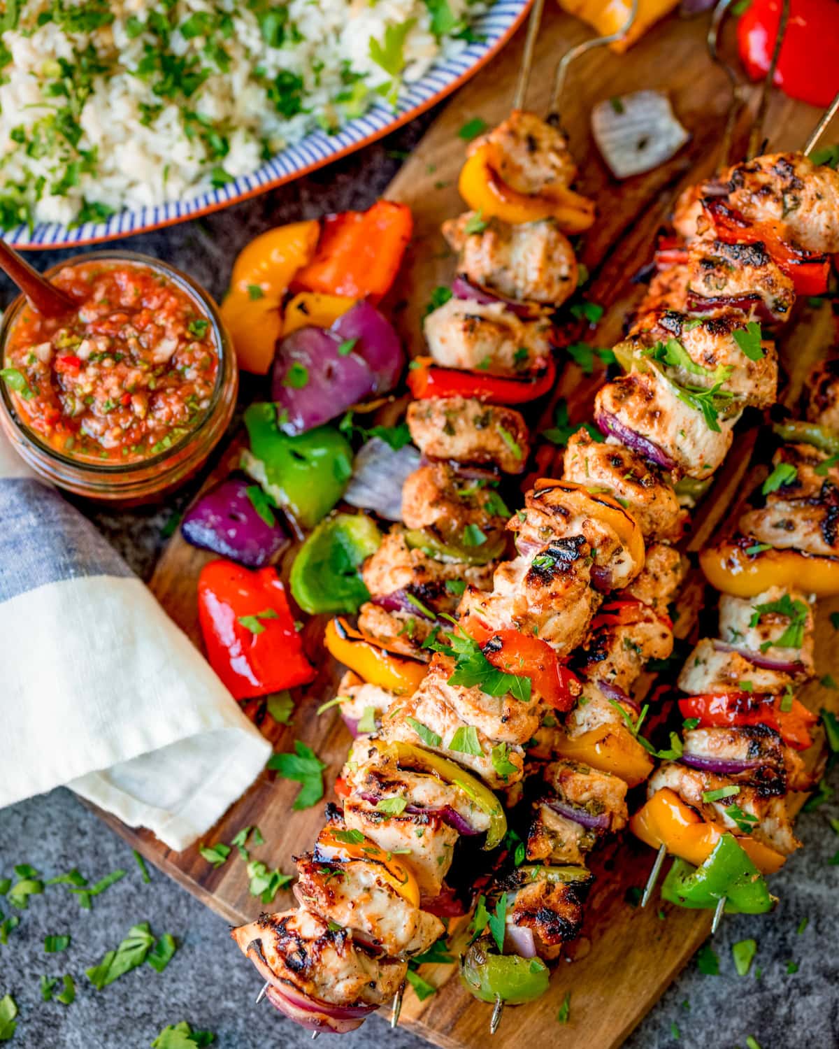 Grilled Mexican style chicken and vegetable skewers on a wooden board, served with a bowl of rice, a jar of salsa, and a serviette.